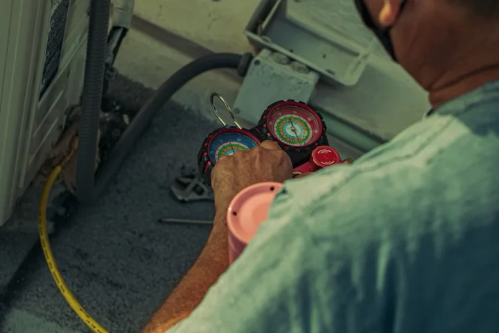 HVAC technician diagnosing an outdoor air-conditioning unit with manifold gauges and service tools.