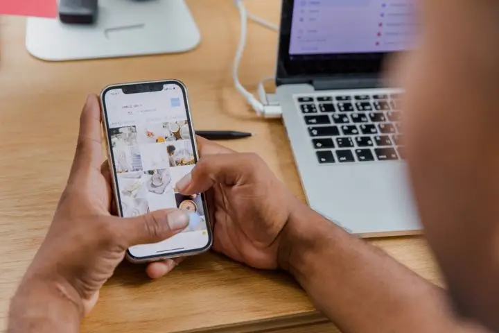 Social media manager reviewing a visual content feed on a smartphone beside a publishing workstation.