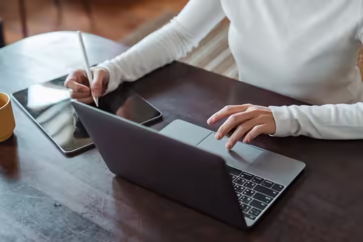 From above of crop anonymous businesswoman in white sweater working at table using tablet with stylus and typing on laptop