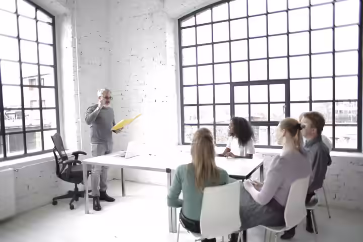Side view of focused multiethnic colleagues listening middle aged speaker while gathering at table in spacious workspace against wide windows