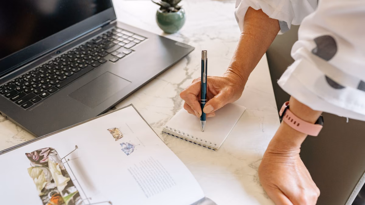 A person writes in a notepad beside a laptop on a desk.