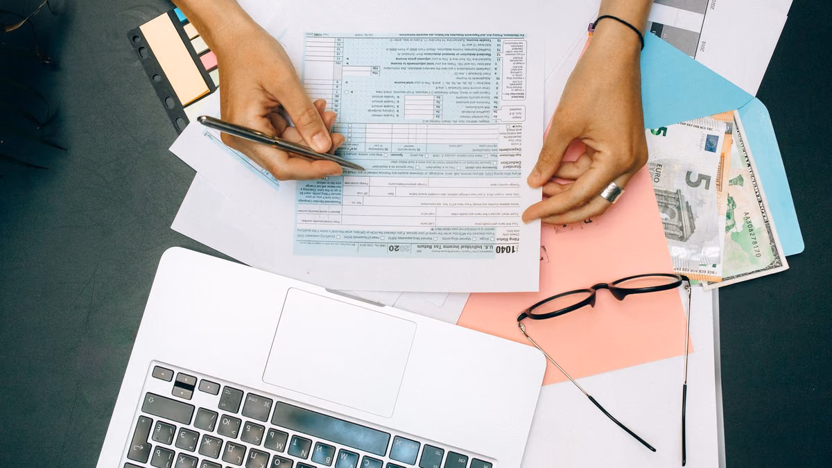 Hands write on financial paperwork beside a laptop and calculator.